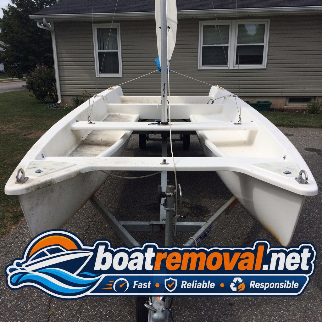 Small catamaran sailboat with faded hulls resting on a trailer beside a house